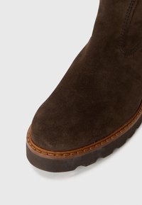 Hnědá semišová kotníková bota s kulatou špičkou, kontrastním prošitím okraje a silnou gumovou podrážkou pro lepší přilnavost. Hladká textura s minimalistickým designem.