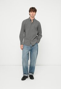 Kockás szürke mintájú gombos ing, hosszú ujjú, világoskék bő szabású farmerrel és fekete cipővel, egyszínű háttér előtt.