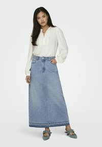 Blouse blanche légère et texturée avec un col volanté, associée à une longue jupe en denim bleue avec un ourlet effiloché et deux poches avant.