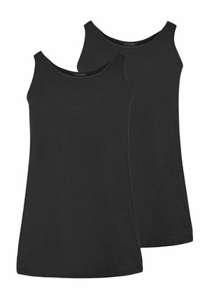 Femme blonde portant un haut noir, une veste en cuir marron foncé drapée sur les épaules, un jean large bleu et des baskets noires, debout contre un mur blanc.