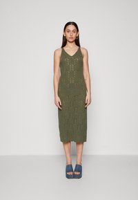 Robe tricotée vert olive avec un col en V, présentant un motif texturé, longueur genou et ourlet festonné. Associée à des chaussures plates-formes bleues.