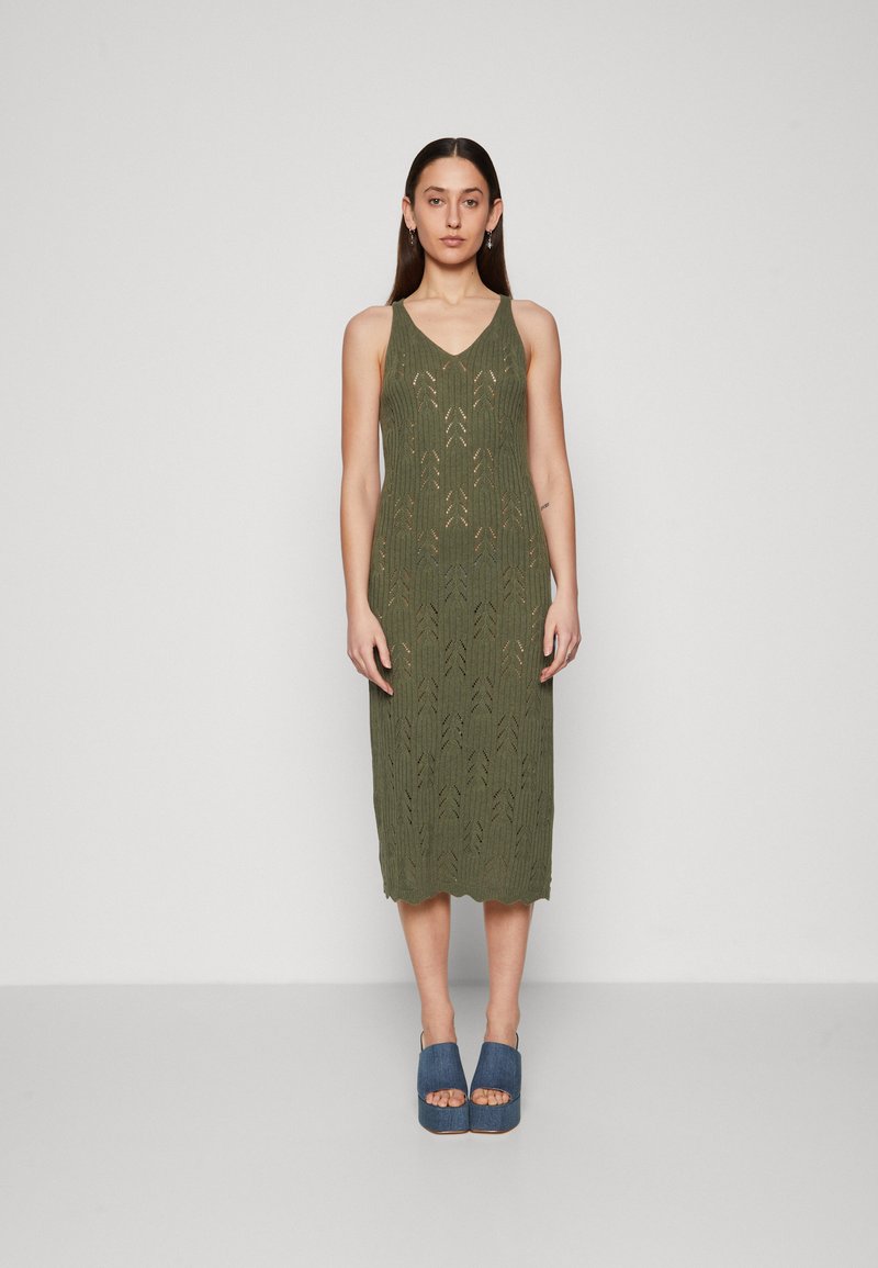 Robe tricotée vert olive avec un col en V, présentant un motif texturé, longueur genou et ourlet festonné. Associée à des chaussures plates-formes bleues.