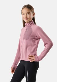 Joven mujer con chaqueta polar rosa con cremallera y leggings negros, de pie con una mano en la cadera, sonriendo ligeramente.