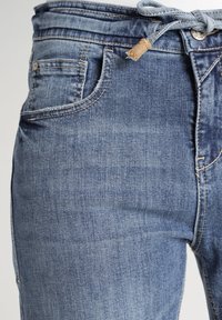 Farkut haalistuneessa sinisessä värissä, jossa on naru vyötäröllä. Farkuissa on etutaskut ja ompelusyksityiskohtia saumoissa.