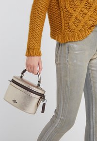 Okrągła, metaliczna złota torba o teksturowanej powierzchni, z czarnym zamkiem błyskawicznym i krótkim uchwytem, trzymana w ręce w musztardowym swetrze z splotem warkoczowym.