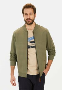 Olijfgroene bomberjack met een gequilt voorkant, geribde manchetten en kraag. Draagt over een beige shirt met een blauw grafisch ontwerp.