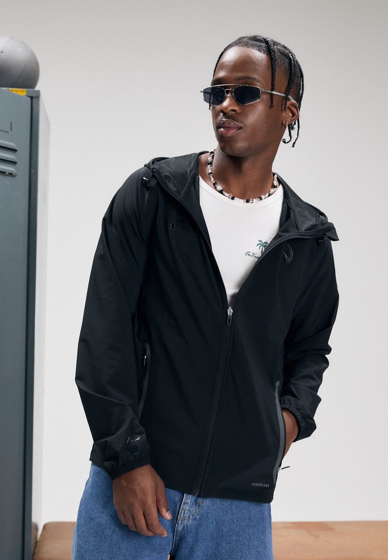 Zwarte lichtgewicht jas met een ritssluiting aan de voorkant, verstelbare capuchon en elastische manchetten. Draagt over een wit overhemd en gecombineerd met blauwe jeans.