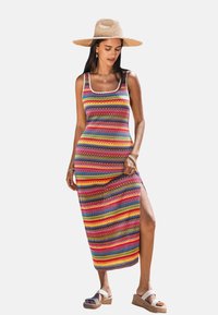 Colorful, striped maxi dress with a scoop neckline, side slit, and knitted texture. Worn with a wide-brimmed straw hat and wedge sandals.