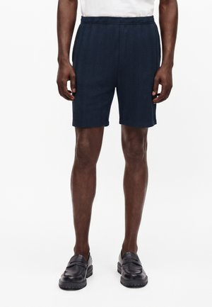 Homme portant un short bleu marine à motif chevrons, des mocassins noirs et une chemise blanche, debout devant un fond blanc uni.