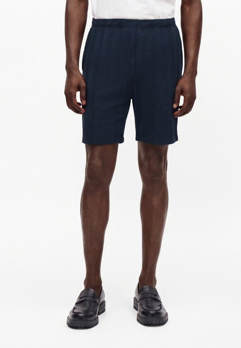 Homme portant un short bleu marine à motif chevrons, des mocassins noirs et une chemise blanche, debout devant un fond blanc uni.