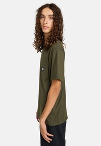 Olive green cotton T-shirt with a round neckline, short sleeves, front pocket, and a black logo patch on the left side.