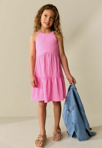 Roza teksturirana poletna obleka s stopničasto spodnjo obrobo, ki jo nosi otrok, ki drži jeans jakno. Obleka ima tanke naramnice in sproščen kroj. Videz dopolnjujejo sandali.