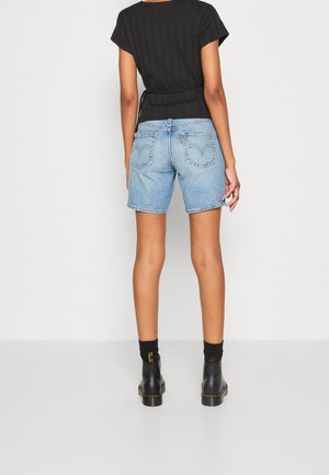 Woman standing facing away wearing a black short-sleeve top, light blue denim shorts, black socks, and black ankle boots.