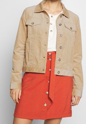 Vrouw met een beige ribfluwelen jas, een wit topje met knopen en een rode rok met knopen aan de voorkant en een strikceintuur, vanaf de schouders tot aan de dijen zichtbaar.