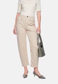 Beige Cordhosen mit geradem Bein, zwei Vorder Taschen und schwarzen Knöpfen, kombiniert mit einem cremefarbenen Oberteil und einer olivgrünen Tasche. Spitz zulaufende Absätze.