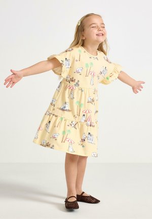 Nuori tyttö keltaisessa mekossa, jossa on piirroshahmoja, ojentaa käsivartensa, sulkee silmänsä ja hymyilee, seisoo yksinkertaisella vaalealla taustalla.