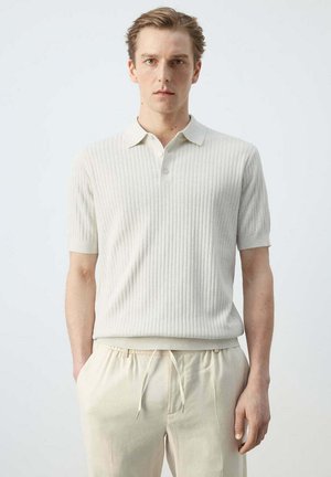 Joven de pie, vistiendo una camiseta polo blanca de manga corta con efecto acanalado y pantalones crema con cordón frente a un fondo liso y claro.