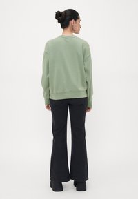 Zelena bombažna majica z udobnim krojem, spušenimi rameni in rebrastim robom, v kombinaciji z razširjenimi črnimi kavbojkami in masivnim črnimi čevlji.