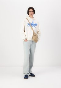 Sudadera blanca con gráfico azul, bolso crossbody beige, vaqueros de corte recto azules claros y zapatillas navy. Diseño sencillo con un ajuste relajado y textura suave.