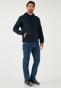 Felpa blu marina con tasca frontale, cappuccio con coulisse e polsini a coste, abbinata a jeans blu e scarpe nere con dettagli grigi.