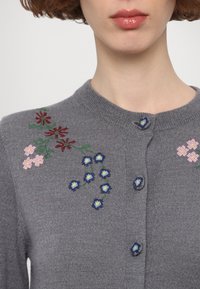 Szürke kötött kardigán virágos hímzéssel rózsaszínben, pirosban és kékben. Kerek nyakkivágással és gombos zárással rendelkezik. Puha tapintású.