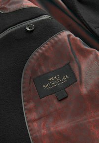 Must mustri kabat mustri külg, millel on punane vooder, millel on äärekese etikett, millel on kirjas "NEXT SIGNATURE PREMIUM TAILORING."