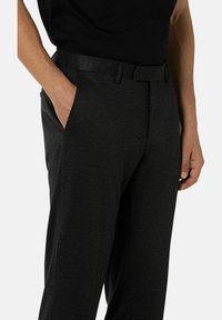 Un homme portant un pantalon noir texturé et une chemise noire, avec une main dans sa poche, devant un fond uni.