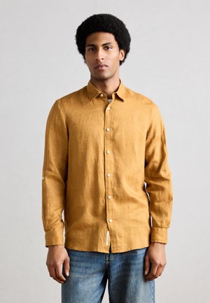 Mladý muž s afrom, oblečený v horčicovo žltej košeli na gombíky s dlhým rukávom a modrých džínsoch, stojaci pred jednoduchým svetlým pozadím.