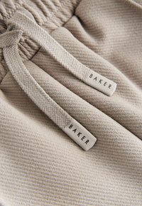 Hellbeiges Gewebe mit einer strukturierten Musterung, versehen mit gewebten Kordeln und einem schwarzen Etikett mit der Aufschrift "BAKER". Nahaufnahme des Materials und der Details.