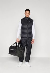 Gilet imbottito nero con cuciture orizzontali, abbinato a pantaloni da ginnastica neri e sneakers bianche. Tiene in mano una borsa da viaggio nera con logo.