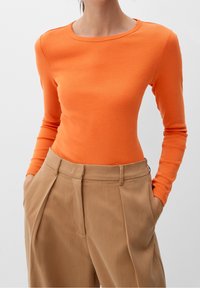 Maglia arancione a maniche lunghe con scollatura rotonda, abbinata a pantaloni plissettati beige a vita alta. Tessuto di cotone morbido.