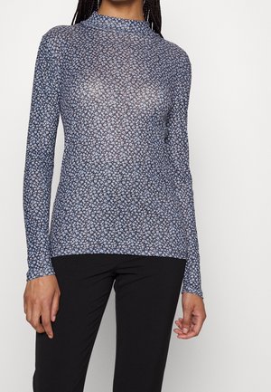 Femme portant un haut bleu marine ajusté à manches longues avec un petit imprimé floral blanc et un pantalon noir, debout devant un fond clair uni.