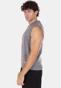 Giovane uomo con capelli ricci che indossa un gilet a maglia grigio senza maniche e pantaloni neri, in piedi di lato su uno sfondo chiaro e uniforme.