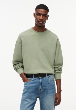 Mężczyzna ubrany w szałwiowozielony, teksturowany sweter z długim rękawem, włożony do jasnoniebieskich dżinsów z czarnym paskiem, pozujący z jedną ręką w kieszeni.