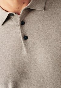 Polo shirt lavorato a maglia beige con tessuto a trama, dotato di colletto e tre bottoni neri. La vista ravvicinata si concentra sulla patta dei bottoni.