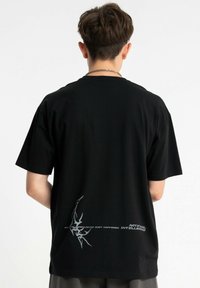 Czarny bawełniany t-shirt z krótkim rękawem, luźnym krojem i srebrnym graficznym wzorem na plecach z tekstem i abstrakcyjnymi akcentami.