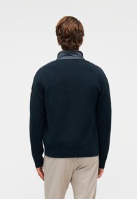 Σκούρο μπλε πλεκτό πουλόβερ με υφή, που διαθέτει αντίθετο γιακά και λεπτομέρεια με σήμα στον αριστερό ώμο.