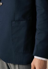 Marineblazer mit Nahaufnahme der Seitentasche, glatter Textur und sichtbaren, kontrastierenden, hellblauen Hemdmanschetten darunter.