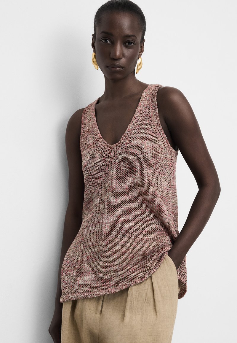 Woman wearing sleeveless pink and beige knit top with V-neck and beige pants, standing with one hand in pocket against plain background.