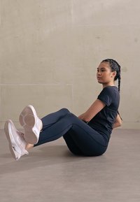 Fekete sportpóló és tengerészkék leggings viselése egy személy által, aki éppen tornagyakorlatot végez, világos színű sportcipővel és fonott hajjal. Simább, modern háttér.