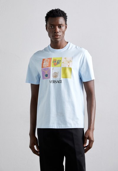 Camiseta de algodón azul claro con diseños gráficos en cuadrados, mostrando motivos de joyería, con el texto "VERSACE" debajo de los gráficos.
