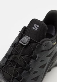 Černá atletická obuv s texturovaným svrškem, nastavitelným systémem tkaniček a zesílenou špičkou. Jemné logo na jazyku a boku.