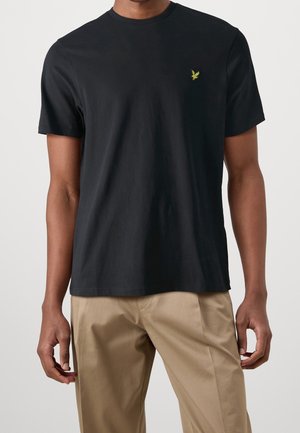 Schwarzes Baumwoll-T-Shirt mit rundem Ausschnitt, kurzen Ärmeln und einem kleinen gelben Stick-Logo auf der linken Brust. Kombiniert mit beiger Hose.