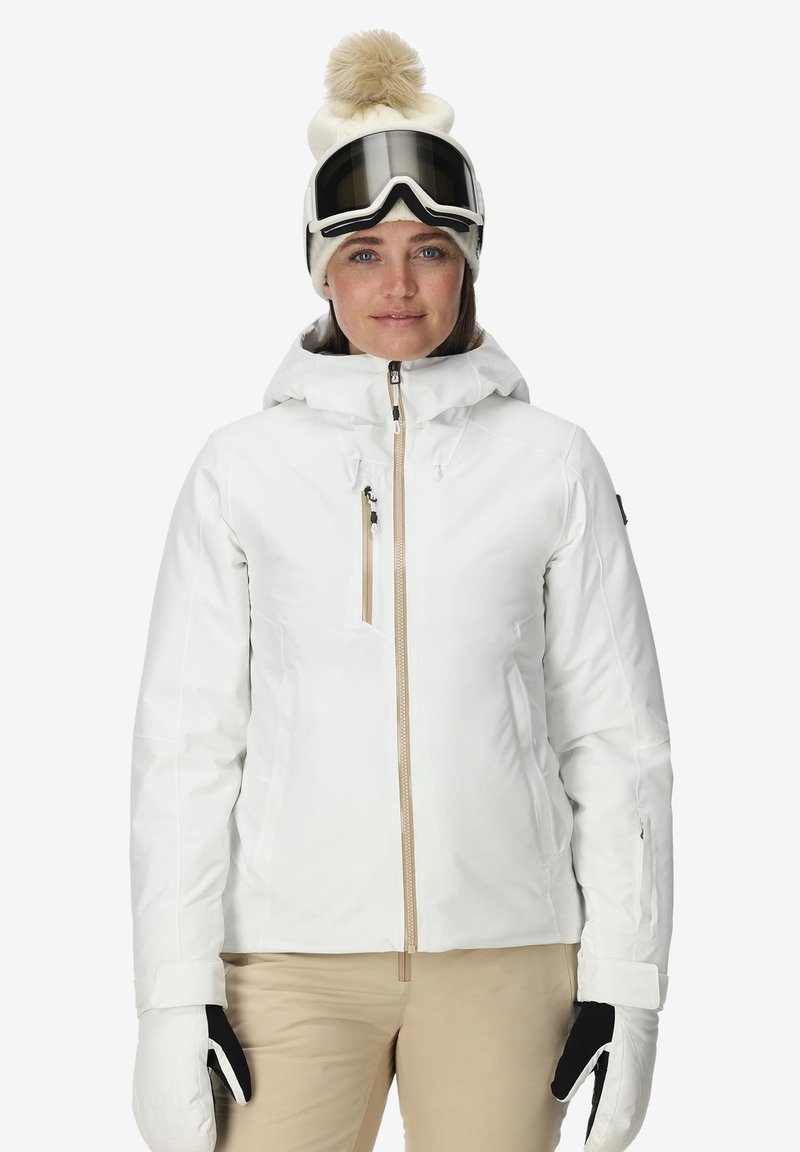 Vrouw in een witte winterjas, beige broek, witte beanie met pompon, skibril op het voorhoofd en witte handschoenen met zwarte palmen.