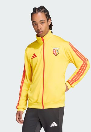 Giovane uomo con capelli intrecciati che indossa una giacca gialla e rossa del Racing Club de Lens e pantaloni neri Adidas con le mani parzialmente nelle tasche.