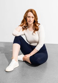 Maglietta a maniche lunghe beige chiaro, leggings a vita alta blu navy e sneaker bianche. Modella seduta su un pavimento grigio, mentre mostra un abbigliamento sportivo casual.