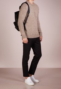 Pull beige en tricot, pantalon noir et baskets blanches ; présente des poignets et un ourlet côtelés, avec un sac à dos noir doté de bretelles réglables.