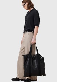 Svart läder tote-väska med tygremmar, stor rektangulär form, slät yta och detalj med dragkedjeficka, hålls av en person.