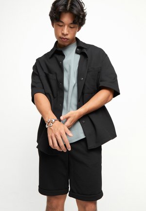 Chemise noire à manches courtes avec boutons sur un t-shirt bleu clair, associée à un short noir. Bracelet en argent notable au poignet.