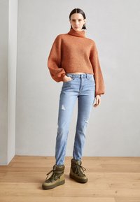 Pull en maille côtelée couleur rouille avec col roulé à manches ballon, associé à un jean droit délavé bleu clair et des bottines vertes.